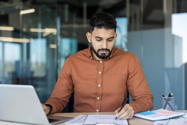 Genç adam masasında çalışıyor, belgeleri analiz ediyor, formları dolduruyor ve modern ofis ortamında dizüstü bilgisayar kullanıyor. Konsantrasyon ve iş verimliliğini temsil ediyor.