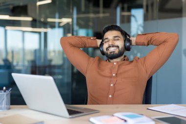 Kulaklık takan ve elleri başının arkasına yaslanan genç adam, müzikten zevk alıyor ve modern ofis ortamında işe ara verirken tatmin oluyor.
