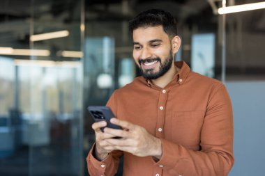 Gülümseyen Hintli adam internette geziniyor ya da cep telefonuyla mesajlaşıyor, çağdaş cam duvarlı ofis ortamında duruyor, ağ, iletişim ve dijital bağlantıyı temsil ediyor.