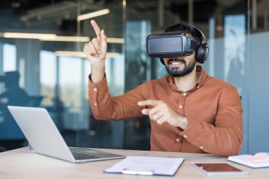 VR kulaklık ve kulaklık takan, gülümseyen, sanal elementlerle iletişim kuran, modern bir ofiste geleceğin teknolojisi ve yeniliği üzerinde çalışan bir adam.