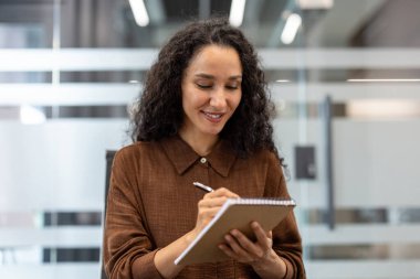 Profesyonel bir iş kadını, aktif olarak bilgi ve fikirleri bir kalemle yazarken gülümsüyor, çağdaş bir ofis ortamında duruyor.