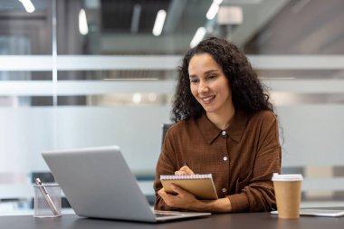 Kıvırcık saçlı, gülümseyen ve notlar yazan bir iş kadını, çevrimiçi bir web sitesine katılırken veya modern bir ofiste dizüstü bilgisayarda buluşurken