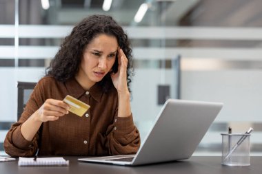 Kredi kartı tutan ve dizüstü bilgisayara bakan genç bir iş kadını, bir ofis ortamında çevrimiçi aktarım yaparken mali sorunlarla karşı karşıya