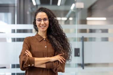 Genç bir kadın gülümsüyor ve kameraya bakıyor, cam bölmelerle modern bir ofiste duruyor ve ışığı yansıtıyor, profesyonelliği, başarıyı ve liderliği temsil ediyor.