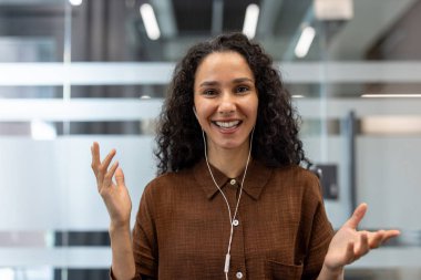 Elleriyle gülümseyen, el kol hareketi yapan, kulaklık takan bir kadın, çağdaş bir ofis ortamından sanal bir toplantıya ya da video görüşmesine katılıyor.