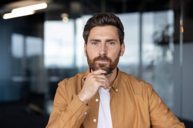 Ciddi derecede iyi hazırlanmış profesyonel bir adamın bir fikri var, çenesinin altında bir kalem tutarken kameraya bakıyor, çağdaş bir iş ortamında strateji planlıyor.