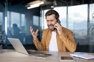 Ofisteki girişimci akıllı telefona bağırıyor, dizüstü bilgisayardaki gergin telefon görüşmesi sırasında sinirleniyor ve stresli, baskı altında öfke, endişe ve işyeri çatışmasını gösteriyor