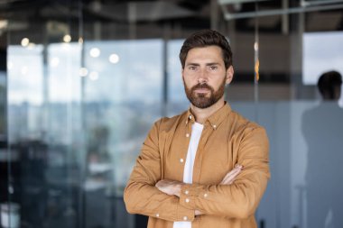 Genç sakallı adam kollarını kavuşturarak poz veriyor. Modern bir ofiste liderlik ve başarıyı temsil ederken kendine güvenini ve profesyonelliğini gösteriyor.