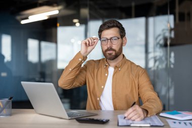 Modern bir ofiste kendine güveni tam bir işadamı masa başında dizüstü bilgisayar kullanarak oturur, notlar yazar ve kameraya bakarken gözlükleri ayarlar, odaklanır ve profesyonelce