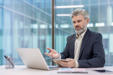 Yetişkin bir iş adamı, ofisinden uzaktan çekilmiş bir video görüşmesinde elinde bir defter, odaklanmış ve sanal bir toplantıyla meşgul bir şekilde el kol hareketi yapıyor.