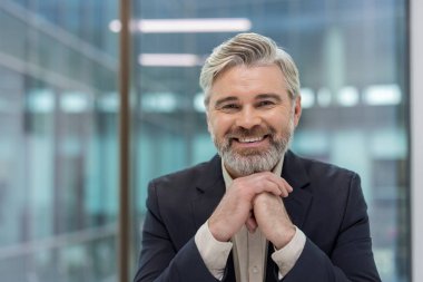 Deneyimli, olgun bir iş adamı kameraya gülümseyip dostça ve kendinden emin bir ifade sergilerken elleri çağdaş bir ofis ortamında sıkıca kenetlenir.