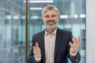Gri sakallı, takım elbiseli, kendine güvenen bir iş adamı modern bir ofiste alkışlıyor ve gülümsüyor, başarıyı kutluyor ve onaylıyor ve mesleki memnuniyet gösteriyor.
