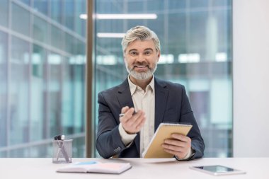Deneyimli bir işadamı modern bir ofiste masa başında oturmuş elinde bir defter ve kalem tutarken kameraya güvenle bakıyor profesyonelliği ve başarılı liderliği somutlaştırıyor.