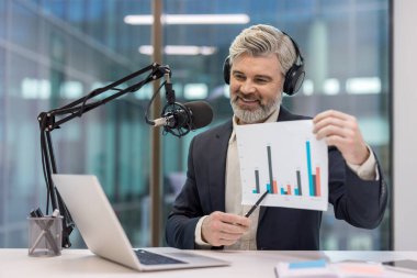 Olgun bir adam online web sitesi ya da podcast 'i kaydediyor, baro tablosu kullanarak finansal verileri açıklarken bir masada dizüstü bilgisayar ve stüdyo mikrofonuyla oturuyor.