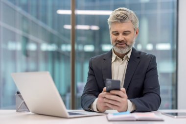 Olgun iş adamı iş sırasında gülümsüyor ve akıllı telefonuyla meşgul oluyor, şirket ofis ortamında dijital iletişim ve teknolojiden faydalanıyor.