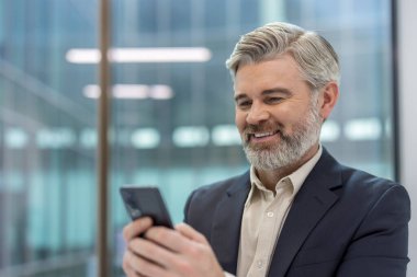 Olgun iş adamı gülümsüyor, takım elbise giyiyor, ofis ortamında akıllı telefon kullanıyor, modern iletişim, teknoloji ve profesyonel ağ kavramlarını temsil ediyor.