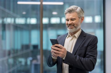 Modern bir ofiste akıllı telefonunu kontrol ederken gülümseyen gri sakallı olgun bir işadamı profesyonel ağ, dijital iletişim, liderlik ve kendinden emin girişimcilik sunuyor.