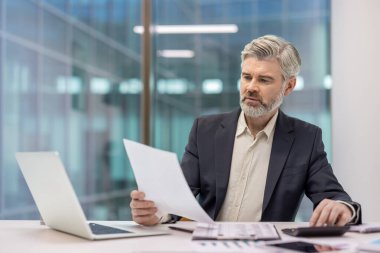 Olgun iş adamı hesap makinesiyle çalışıyor, mali raporları analiz ediyor ve elinde muhasebe, finans ve kurumsal yönetim kavramlarını tasvir eden bir belge tutuyor.