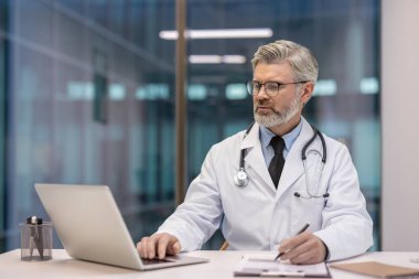 Beyaz önlük ve steteskop giyen olgun bir erkek doktor, bir masada oturuyor, bir dizüstü bilgisayarda yazıyor ve modern bir ofis veya klinikte not alıyor.