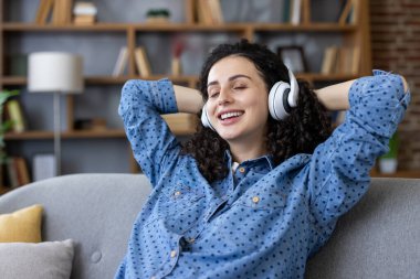 Kapalı gözlü ve mutlu bir ifadeyle beyaz kulaklıkla müziğin tadını çıkaran genç bir kadın evinde gri bir koltukta rahatça oturup, rahatça dinleniyor ve sakin bir şekilde dinleniyor.
