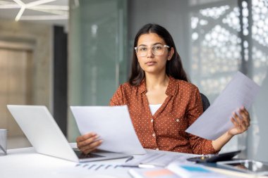 Genç Hintli iş kadını masasında çalışıyor, elinde bir dizüstü bilgisayar ve yakınlardaki çeşitli ofis malzemeleriyle kameraya bakarken evrakları tutuyor ve inceliyor.