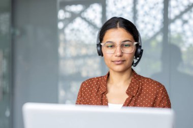 Kulaklık takan ve dizüstü bilgisayarda çalışan genç bir kadın, müşteri desteği ya da özel çağrı merkezi operatörü olarak teknik destek sağlıyor