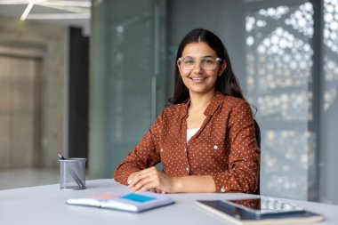 Genç Hintli şirket çalışanı gözlüklü ve noktalı tişörtlü. Çağdaş bir ofis ortamında defter ve tabletle bir masada otururken özgüvenle gülümsüyor.