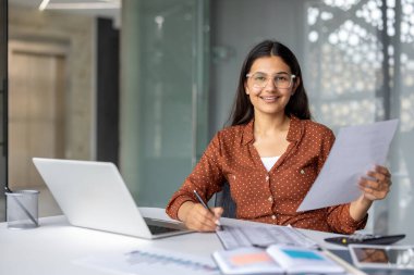 Genç Hintli kadın muhasebeci gülümsüyor, elinde belgeler tutuyor ve notlar yazıyor. Modern bir ofiste, bilgisayarında çalışırken, finans, iş ve başarıyı temsil ediyor.