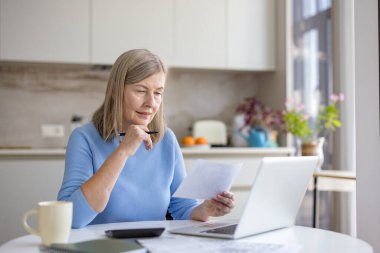 Finansal belgeleri ve faturaları analiz eden olgun bir kadın, mutfak masasındaki harcamaları hesaplarken aynı zamanda emeklilik birikimlerini hesaplamak ve planlamak için bir dizüstü bilgisayar kullanıyor.