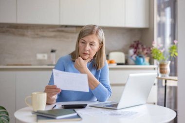 Modern mutfakta kıdemli bir kadın masalarda fatura ve harcamaları hesaplıyor, evrak işlerini ve dizüstü bilgisayarları ciddi ve endişeli bir ifadeyle gözden geçirip bütçe ve mali planlama yaparken