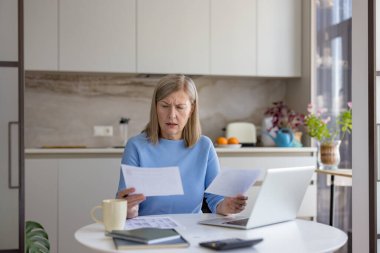 Üst düzey bir kadın, evde kişisel mali durumlarını yönetirken ve aylık faturaları hesaplarken stresli ve endişeli hissediyor, masadaki bilgisayar ve hesap makinesiyle kağıt belgelere bakıyor.