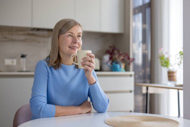 Son sınıf öğrencisi kadın evde dinleniyor. Mutfağında sıcak bir fincan kahve tutarken gülümsüyor. Düşüncelilik ve rahatlıkla dolu huzurlu bir sabahın tadını çıkarıyor.