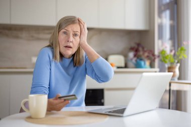 Evde elinde bir kredi kartı ve dizüstü bilgisayar olan yaşlı bir kadın, online alışveriş veya bankacılık girişiminde bulunurken stres ve kafa karışıklığı yaşıyor, olası bir dolandırıcılığa veya dolandırıcılığa tepki gösteriyor