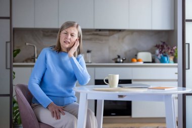 Yaşlı bir kadın, acı veren bir kulak ağrısı çekiyor, yüzünü buruşturan bir ifadeyle elini tutuyor, mutfak masasında oturuyor ve rahatlamaya ihtiyacı var.