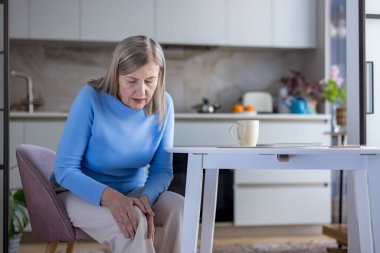 Son sınıf öğrencisi kadın mutfakta oturmuş, ağrılı dizini kavrayıp yaşla ilgili eklem ağrısı ve eklem iltihabı yüzünden rahatsızlığını gösteriyor ve günlük ev hayatını ve hareketliliğini etkiliyor.