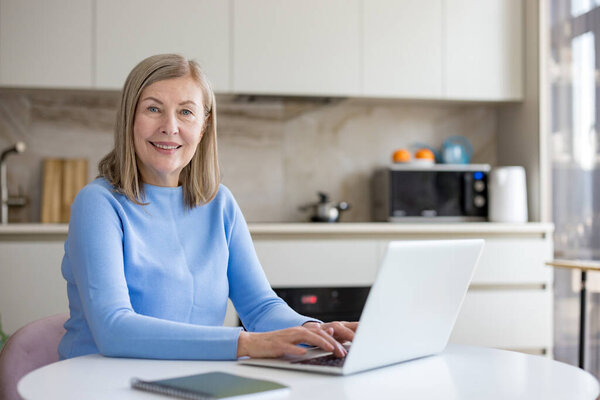 Senior woman smiling while typing on laptop at kitchen table, working remotely and learning online-confident, connected, and enjoying modern digital, freelance lifestyle at home