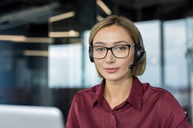 Kulaklık ve gözlük takan profesyonel kadın operatör, kameraya gülümsüyor, modern ofis ortamında müşteri desteği ve tele pazarlama hizmetleri sunmaya hazır.