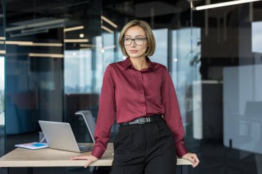 Genç profesyonel iş kadını ofis masasının başında duruyor, modern bir şirket çalışma alanı içinde gelecekteki iş stratejilerini tasarlıyor.