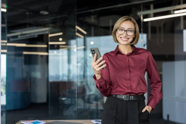 Güncel bir iş yerinde başarılı, iletişim ve bağlantıyı temsil eden, gülümseyen ve elinde akıllı bir telefon tutan kendine güvenen bir iş kadını.