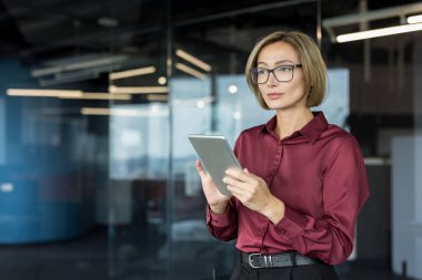 Bardaklı Asyalı iş kadını elinde tablet, modern açık planlı bir ofiste duruyor, liderliği somutlaştırıyor, teknolojik bütünleşme ve profesyonel kararlılığa odaklanıyor.