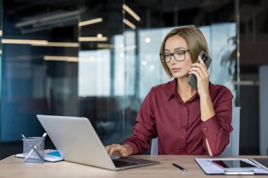 Modern çalışma ofisinde çalışan, cep telefonuyla konuşan ve dizüstü bilgisayar kullanan, uzaktan çalışma, iletişim ve üretkenlik kavramlarını tasvir eden kadın profesyonel