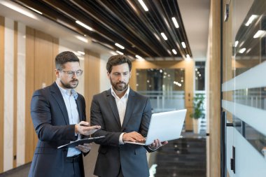İki profesyonel işadamı modern bir şirket koridorunda proje detaylarını tartışıyor, biri dizüstü bilgisayar tutarken, diğeri de takım çalışmasını ve verimliliğini temsil eden bir panoya işaret ediyor.