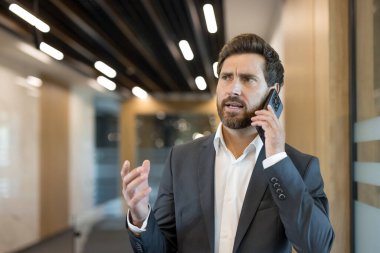 Takım elbiseli ve beyaz gömlekli bir iş adamı cep telefonuyla ciddi bir konuşma yapıyor, eliyle işaret ediyor ve modern bir ofis koridorunda dikilirken endişeli görünüyor.