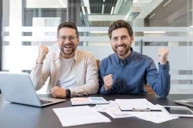 İki neşeli iş adamı bir masada dizüstü bilgisayar ve belgelerle oturuyor, kutlama için yumruklarını kaldırıyorlar, bir hedefe ulaşmak ya da başarılı bir anlaşma yapmak için mutlular.