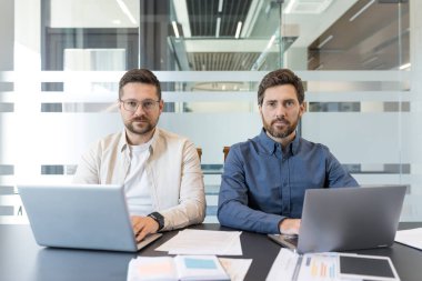 Modern bir ofis masasında oturan iki profesyonel iş adamı, ortak bir şirket toplantısı sırasında doğrudan kameraya bakarken aktif olarak dizüstü bilgisayarları ve kağıtları üzerinde çalışıyorlar.