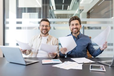 İki neşeli erkek meslektaşı modern bir ofis masasında ellerinde evrak ve kağıtlarla oturmuş başarılı takım çalışmalarını ve bir toplantı sırasında olumlu iş sonuçlarını kutluyorlar.