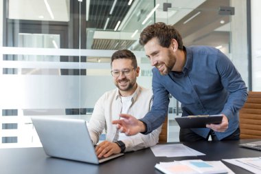Bir dizüstü bilgisayarda birlikte çalışan, bir projeyi tartışan ve çağdaş ofis ortamında verimli bir toplantı sırasında ekranı işaret eden iki yetişkin iş adamı.