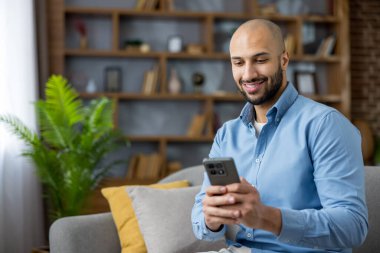 Evdeki bir kanepede gülümseyen ve rahatlatan genç bir adam, akıllı telefonuyla sosyal medyaya göz gezdirirken dijital içeriğe bağlanıyor, modern iletişimi ve eğlenceyi temsil ediyor.