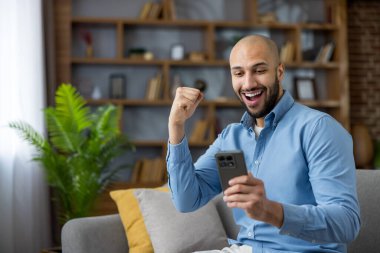 Modern bir oturma odasındaki kanepede oturan genç adam mutlu bir şekilde telefonuna bakar, bir zaferi ya da iyi haberi bir gülümseme ve muzaffer bir heyecanla kutlar.