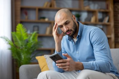 Endişeli yüz ifadesi olan bir adam koltukta oturuyor, akıllı telefonu tutuyor ve ekrana bakıyor, kötü haber aldıktan sonra stresli ve endişeli, modern hayatın gergin bir anı.